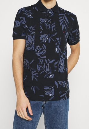 Mann trägt schwarzes Polohemd mit dunkelblauen Blumenmustern und blauen Jeans, steht mit einer Hand in der Tasche.