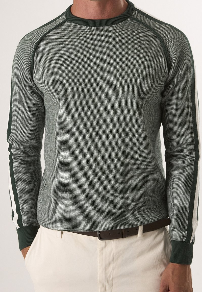 Homme portant un pull vert texturé à manches longues avec des rayures blanches et vert foncé sur les manches, pantalon beige et ceinture marron, mains dans les poches.