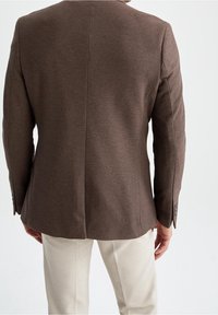 Uomo vestito con un blazer marrone aderente e pantaloni beige chiaro, visto di spalle su uno sfondo bianco uniforme.