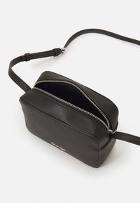 Zwarte leren crossbodytas met een rechthoekige vorm, zilveren rits, smal hengsel en minimalistisch ontwerp. De binnenkant is onbewerkt.