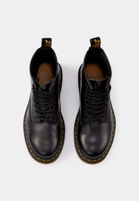 Bottines en cuir noir avec lacets noirs, coutures jaunes, bout rond et une languette. Texture lisse avec une semelle en caoutchouc robuste.