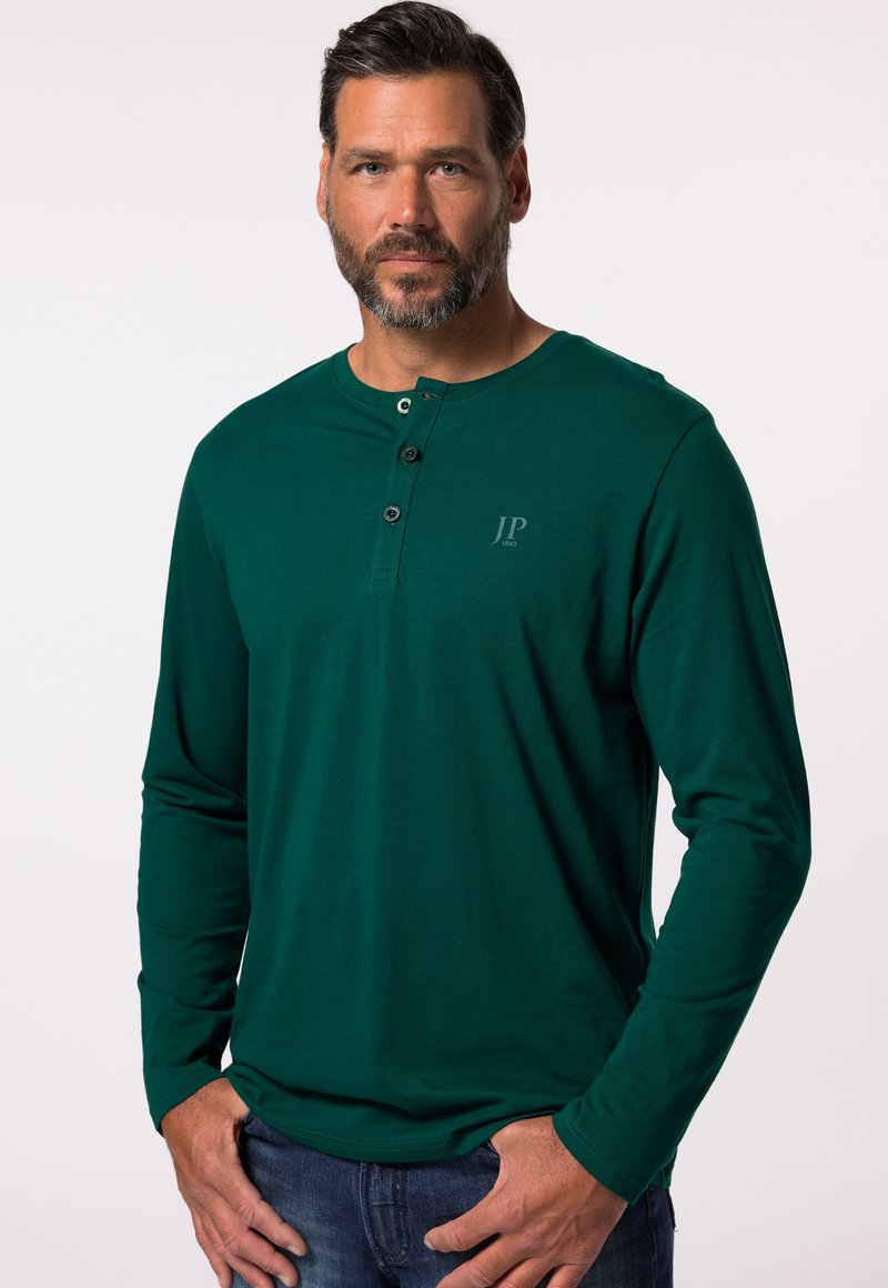 Maglietta henley a maniche lunghe di color verde scuro con tre bottoni sul collo, dotata di un logo discreto sul petto. Tessuto morbido e liscio.