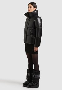 Melnā piepūšamā jaka ar augstu apkakli, spīdīga virsma un priekšējām kabatām, apvienota ar melnu ribotu svārku un melniem sniega zābakiem.