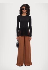 Top negru cu mâneci lungi, cu tiv franjurat, asociat cu pantaloni maro, cu talie înaltă și lățime largă. Modelul ține o geantă de mână mică, neagră.