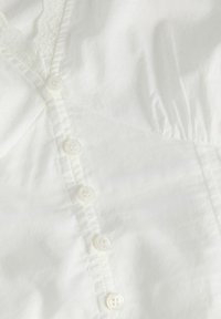 Tissu blanc avec une rangée verticale de six petits boutons translucides et des détails subtils de fronces le long des bords.