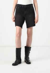 Shorts en denim noir avec un ourlet brut, présentant un design classique à cinq poches et une taille ajustée. Associés à des bottes en cuir noir montantes.