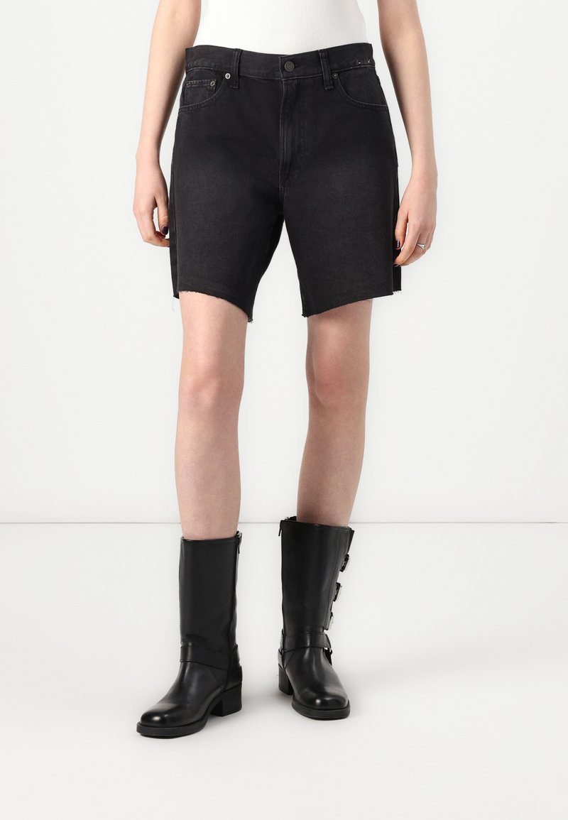 Shorts en denim noir avec un ourlet brut, présentant un design classique à cinq poches et une taille ajustée. Associés à des bottes en cuir noir montantes.