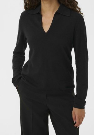 Femme en pull noir à col en V à manches longues avec col et pantalon noir, une main dans la poche, debout devant un fond uni.