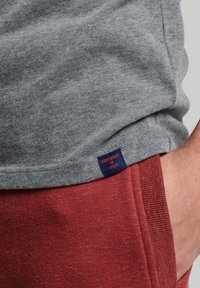 T-shirt en coton gris avec une étiquette en tissu bleu marine à l'ourlet, associé à un pantalon de survêtement rouge avec une taille côtelée et une texture.