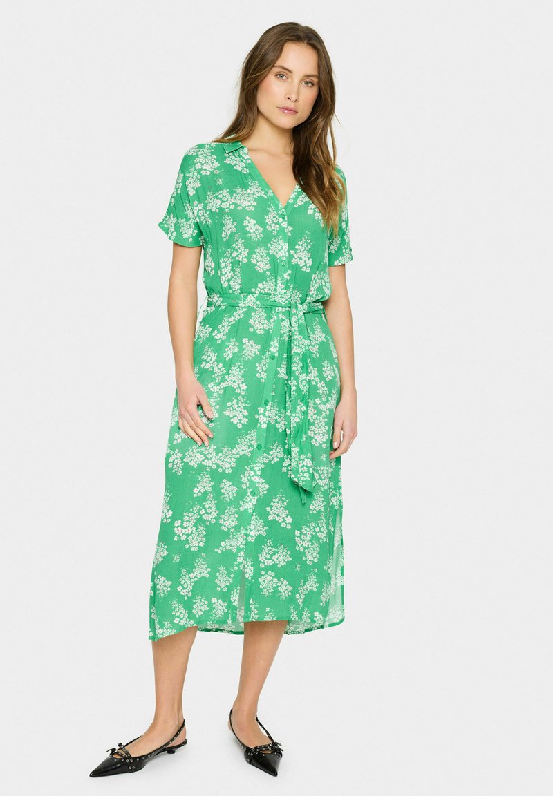 Rochie florală verde cu mâneci scurte, design cu butoni, curea în talie și lungime midi vaporoasă. Pantofii negri cu vârf ascuțit completează ținuta.
