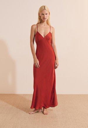 Mujer rubia de pie descalza sobre una alfombra beige, con un vestido rojo sin mangas que llega hasta el suelo y con finas tiras en los hombros.