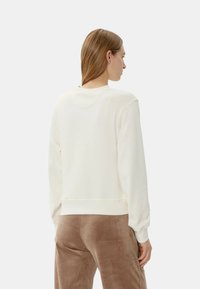 Felpa bianca con cappuccio e polsini a costine, caratterizzata da una vestibilità rilassata e una texture morbida, abbinata a pantaloni marroni in velluto.