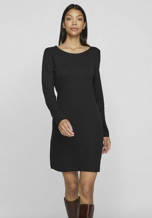 Vestido negro de manga larga con escote redondo, silueta ajustada y tejido suave. El vestido termina por encima de la rodilla, combinado con botas marrón hasta la rodilla.