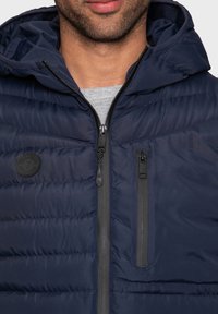Marineblaue Pufferjacke mit Kapuze, ausgestattet mit einer zipbaren Brusttasche, strukturiertem Material und einem runden Logo-Patch auf der oberen linken Seite.