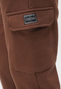 Brune stofbukser med sidelomme med et rektangulært mærke med teksten "PIER ONE." Materialet er blødt med en glat tekstur.