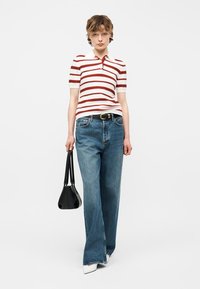 Jeune personne portant un polo à manches courtes rayé blanc et rouge, un jean à jambes larges bleu avec une ceinture noire, des chaussures blanches, et tenant un sac à main noir.