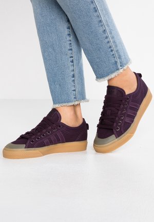 Osoba w fioletowych płóciennych trampkach z gumowymi podeszwami oraz w jasnoniebieskich dżinsach z postrzępionymi nogawkami, stojąca na białej podłodze.