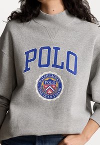 Felpa grigia con collo alto, caratterizzata da grande scritta blu "POLO" e un'emblema circolare con "Ralph Lauren" e elementi decorativi.