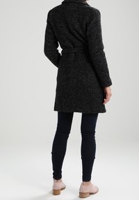 Cappotto in misto lana nero con vita cinturata, maniche lunghe e colletto. Abbinato a jeans scuri e scarpe rosa chiaro con tacco a blocco.