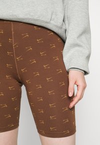 Smeđi atletski šortsevi s ponavljajućim žutim Nike logotipom i tekstualnim uzorkom, izrađeni od mekog, rastezljivog materijala. Uparite s sivim duksericom.