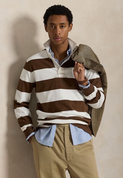 Giovane uomo che indossa una maglia da rugby a righe marrone e bianca sopra una camicia celeste con bottoni, pantaloni beige, che tiene una giacca spina di pesce sulla spalla.