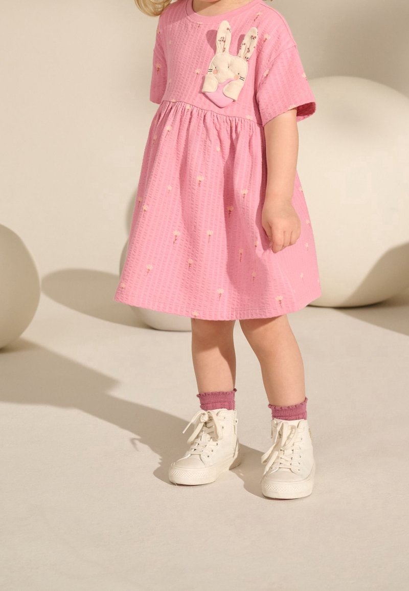 Tout-petit portant une robe rose avec de petits motifs floraux et une poche avec un lapin en peluche, des chaussettes violettes et des baskets blanches montantes.