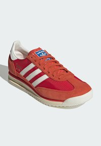 Chaussure de sport rouge et orange avec des rayures blanches, en matériaux de suède et de mesh, semelle beige, bout arrondi et étiquette bleue sur la languette.