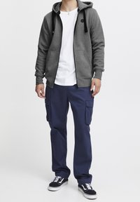 Sudadera gris con cremallera y cordones negros, camiseta blanca debajo, pantalones cargo azul marino y zapatillas negras con detalles blancos.