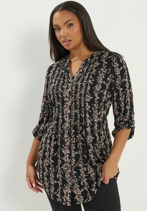 Kvinde iført en sort bluse med lange ærmer og lyserødt blomsterprint, stående mod en ensfarvet lys baggrund, hånd i lommen.