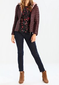 Bruine pufferjack met roze voering, zwarte top met bloemenprint, donkerblauwe jeans en beige enkellaarsjes; getailleerd design met contrasterende kleuren en texturen.