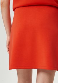 Rode skaterskirt van zachte stof, heeft een uitlopende silhoeet, een gladde textuur en een nette zoom. Handen houden de rok bij de taille vast.
