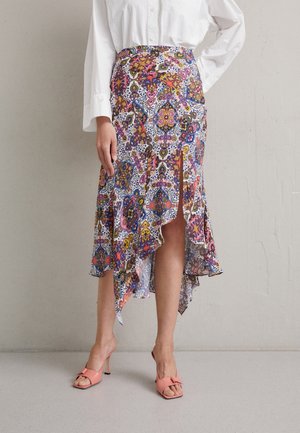 Personne portant une jupe à motif haut-bas avec des motifs floraux et géométriques colorés et des sandales à talons ouvertes roses, debout contre un mur uni.