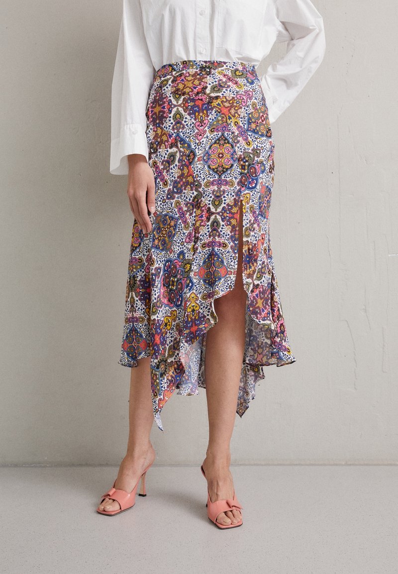 Personne portant une jupe à motif haut-bas avec des motifs floraux et géométriques colorés et des sandales à talons ouvertes roses, debout contre un mur uni.