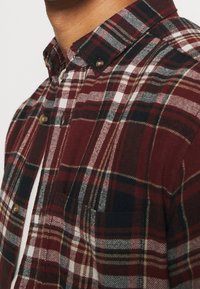 Chemise en flanelle à carreaux rouge et marine avec un col boutonné, boutons marron et une poche poitrine. Texture douce avec un motif à carreaux classique.