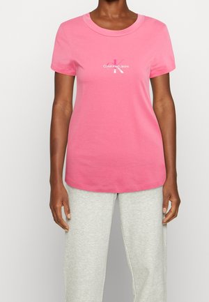 Kobieta ubrana w różowy t-shirt Calvin Klein Jeans i jasnoszare spodnie dresowe, stojąca na tle jednolitego białego tła.