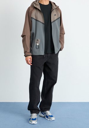 Lätt, vattentät jacka med grå och bruna paneler, dragkedjefickor, justerbar huva och textilmaterial, bärs över en svart skjorta och jeans.