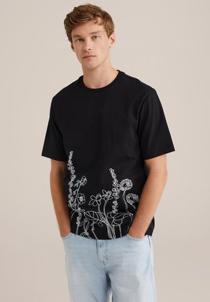 Black cotton t-shirt with short sleeves and a round neck, featuring white floral line drawings at the bottom hem. Light denim jeans.