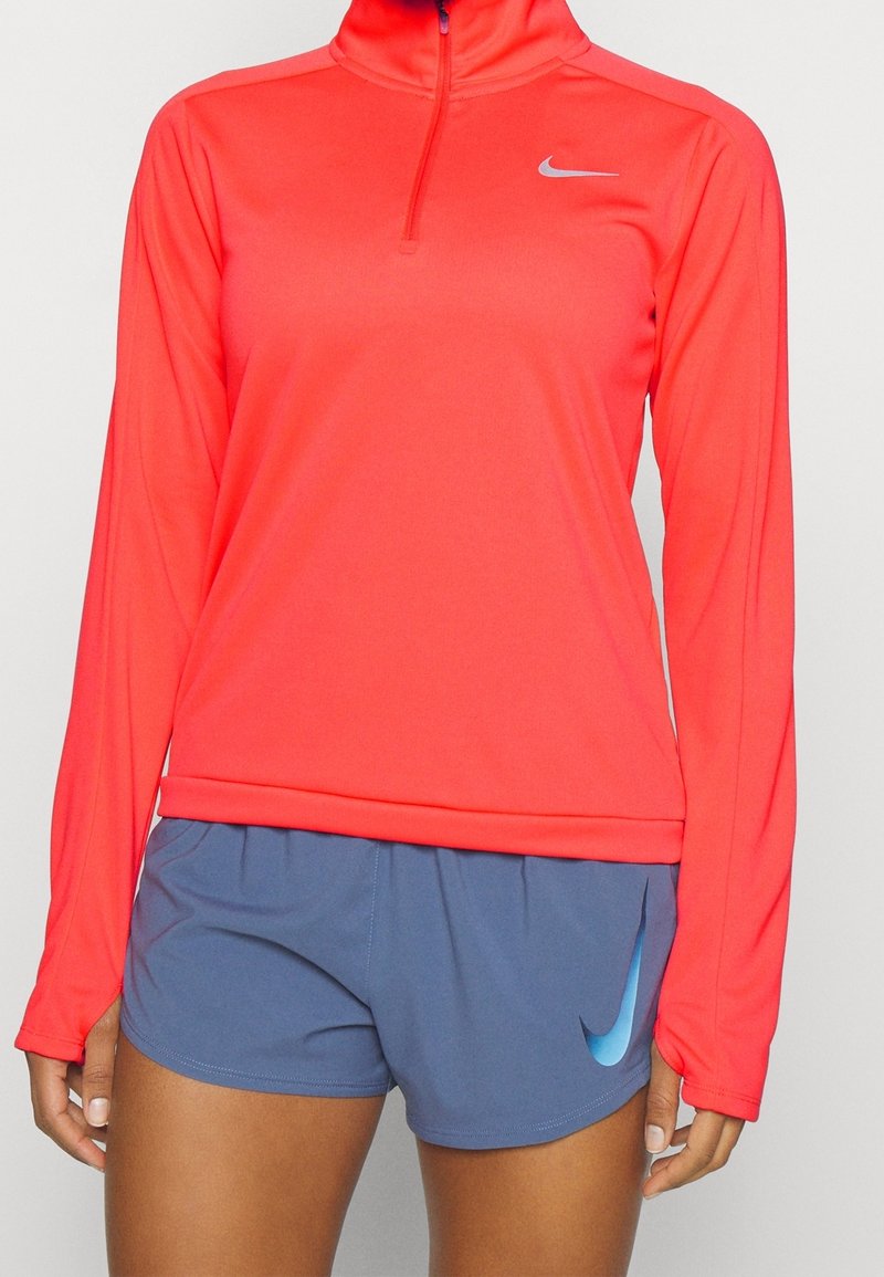 Pull de sport à demi-zip Nike en corail vif, fabriqué en tissu léger, assorti à un short bleu avec un accent logo bleu clair.