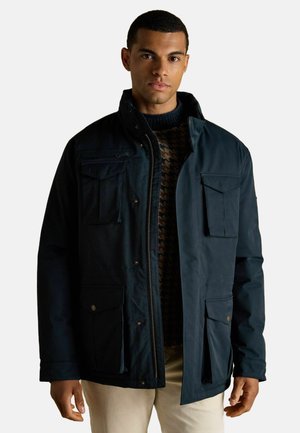 Navy blue waterproof jacket with a zip front, multiple front pockets, and a high collar, layered over a patterned sweater.