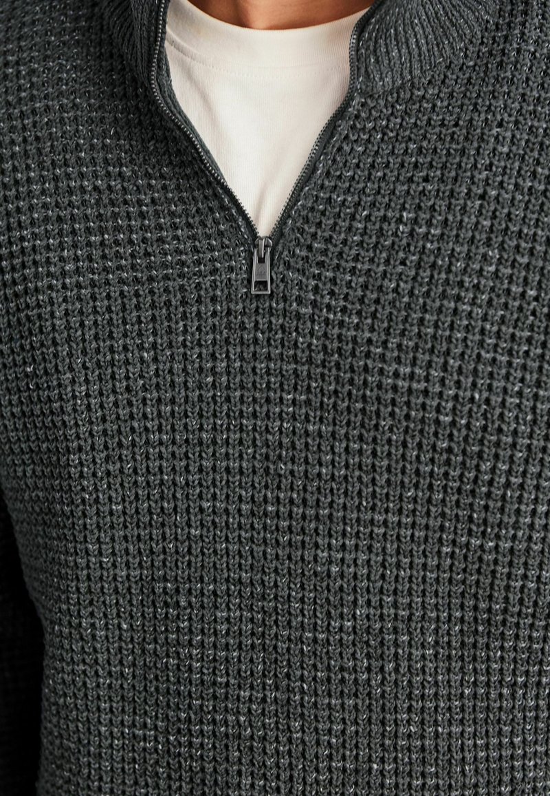 Pulover din tricot gri închis cu guler înalt și un fermoar pe jumătate. Are un model texturat și un strat interior mai deschis ca nuanță, în contrast.