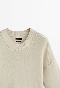 Beżowy sweter dziany z okrągłym dekoltem, prążkowaną fakturą i subtelnymi detalami na szwie ramienia. Wewnątrz widoczna metka.