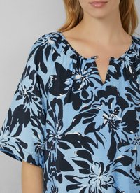 Frau, die eine blaue Bluse mit einem großen schwarz-weißen Blumendruck trägt, mit einem runden Ausschnitt und einem kleinen Knopfverschluss.