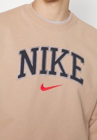 Beige sweatshirt med rundhals. Har "NIKE" i svarta texturerade bokstäver och en röd swoosh-logotyp. Tillverkad av mjukt material med en slät finish.