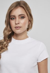 Bílé žebrované tričko s krátkým rukávem a volánkovým výstřihem. Modelka má vlnité vlasy a náušnice ve tvaru kruhu, na neutrálním pozadí.