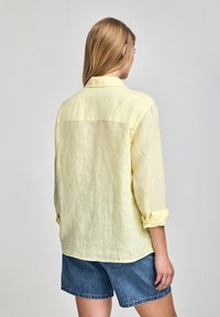 Ljust gult linneskjorta med avslappnad passform, knäppning fram, uppvikta ärmar och rundad nederkant. Bärs tillsammans med jeansshorts.