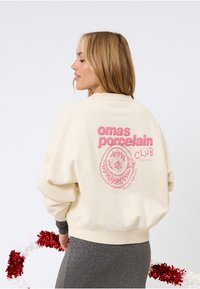 Sweatshirt oversize couleur crème avec un motif rose représentant une tasse à fleurs et le texte "omas porcelain CLUB." Poignets ajustés.