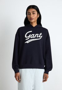 Navy blue oversized hoodie made of soft fabric featuring a large white "Gant" logo with a curved underline and ribbed cuffs and hem.