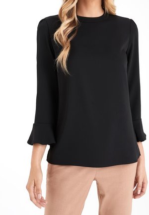 Femme portant un chemisier noir à manches longues avec des poignets à volants et un pantalon beige, avec cheveux blonds ondulés mi-longs visibles.