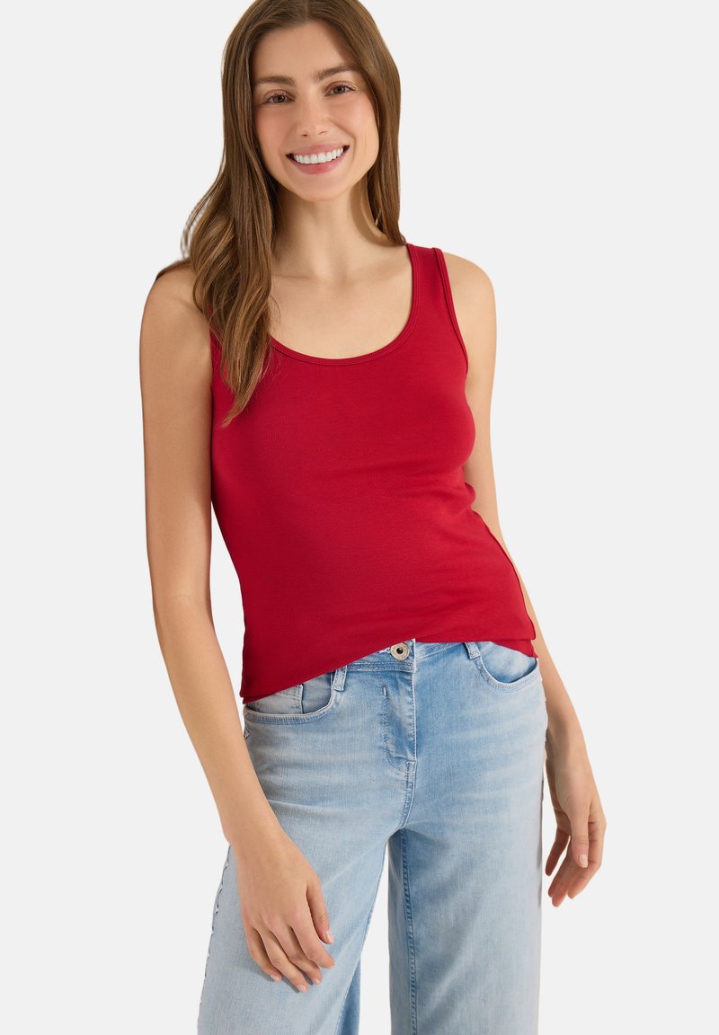 Jeune femme aux longs cheveux bruns souriante, portant un haut sans manches rouge et un jean bleu clair, debout devant un fond uni.