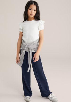 Niña pequeña con cabello largo y oscuro, vistiendo una blusa blanca con mangas acampanadas, pantalones anchos azul marino, un suéter gris atado a la cintura y zapatillas blancas.